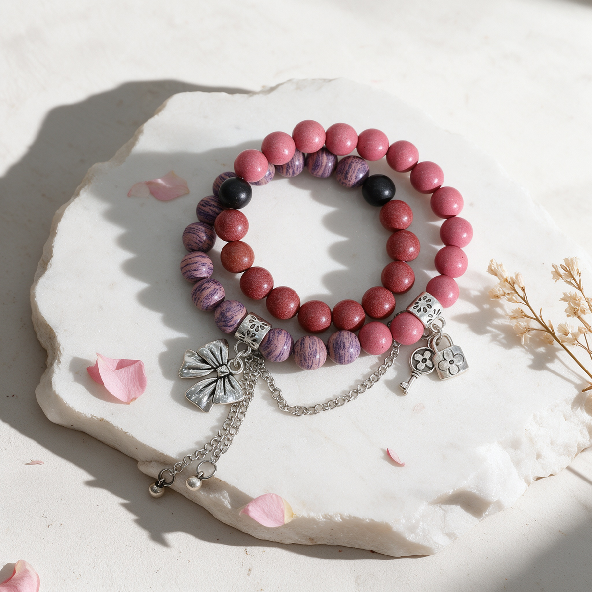 Top view of a double-wrap scented incense bead bracelet with blush pink, wine red, marbled violet and black beads, carved silver spacer beads, bow tassel and lock and key charms resting on a white stone surface